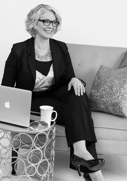 Linda Rey sitting on a couch with laptop and cup of coffee on a small table looking away from camera